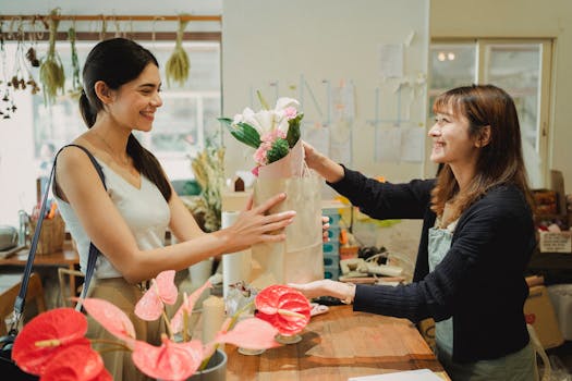 Side view of cheerful florist in apron giving paper bag with bouquets of flowers to smiling customer in floral shop in daytime