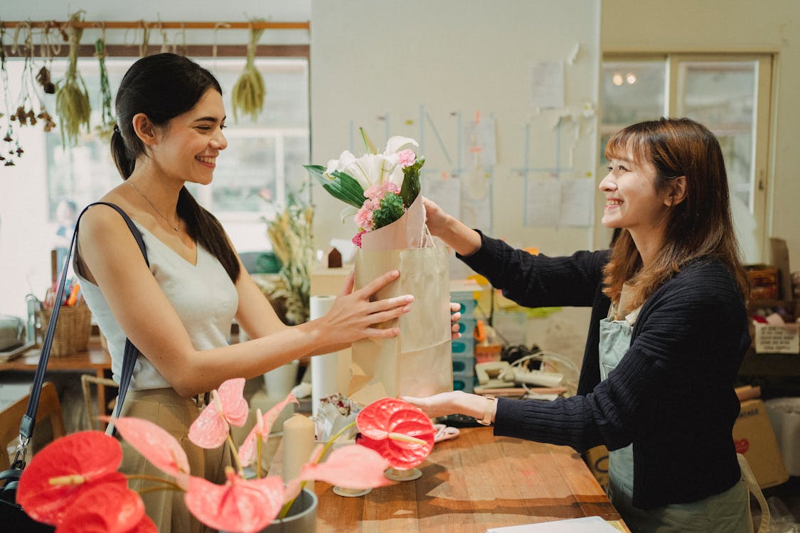 Free Side view of cheerful florist in apron giving paper bag with bouquets of flowers to smiling customer in floral shop in daytime Stock Photo