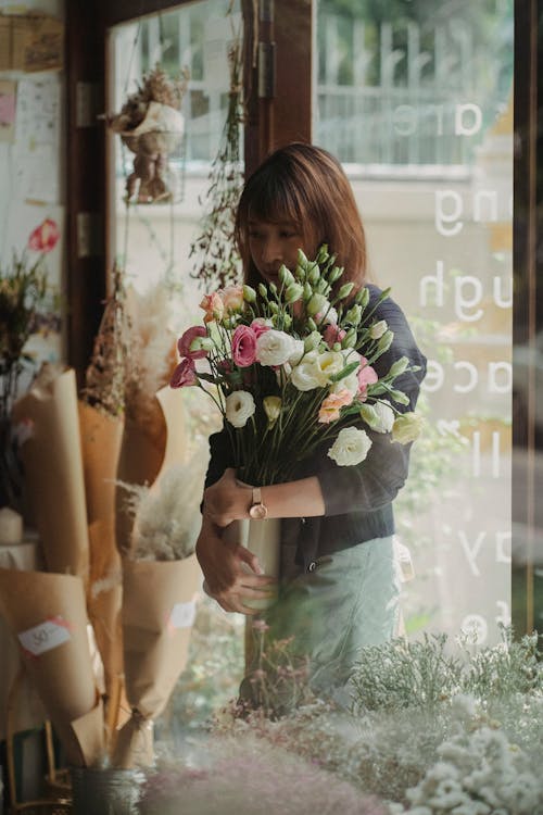 Through glass woman carrying vase with blossom flowers while standing near entrance in shop