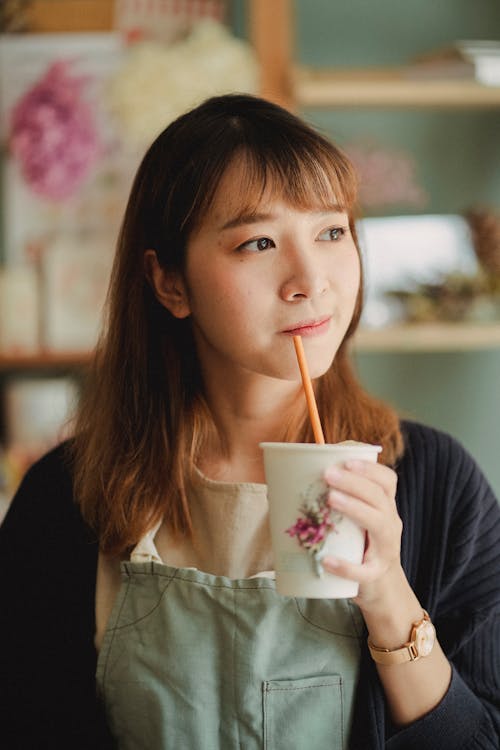 Calm Asian woman in casual clothes looking away enjoying paper cup of delicious coffee with tube