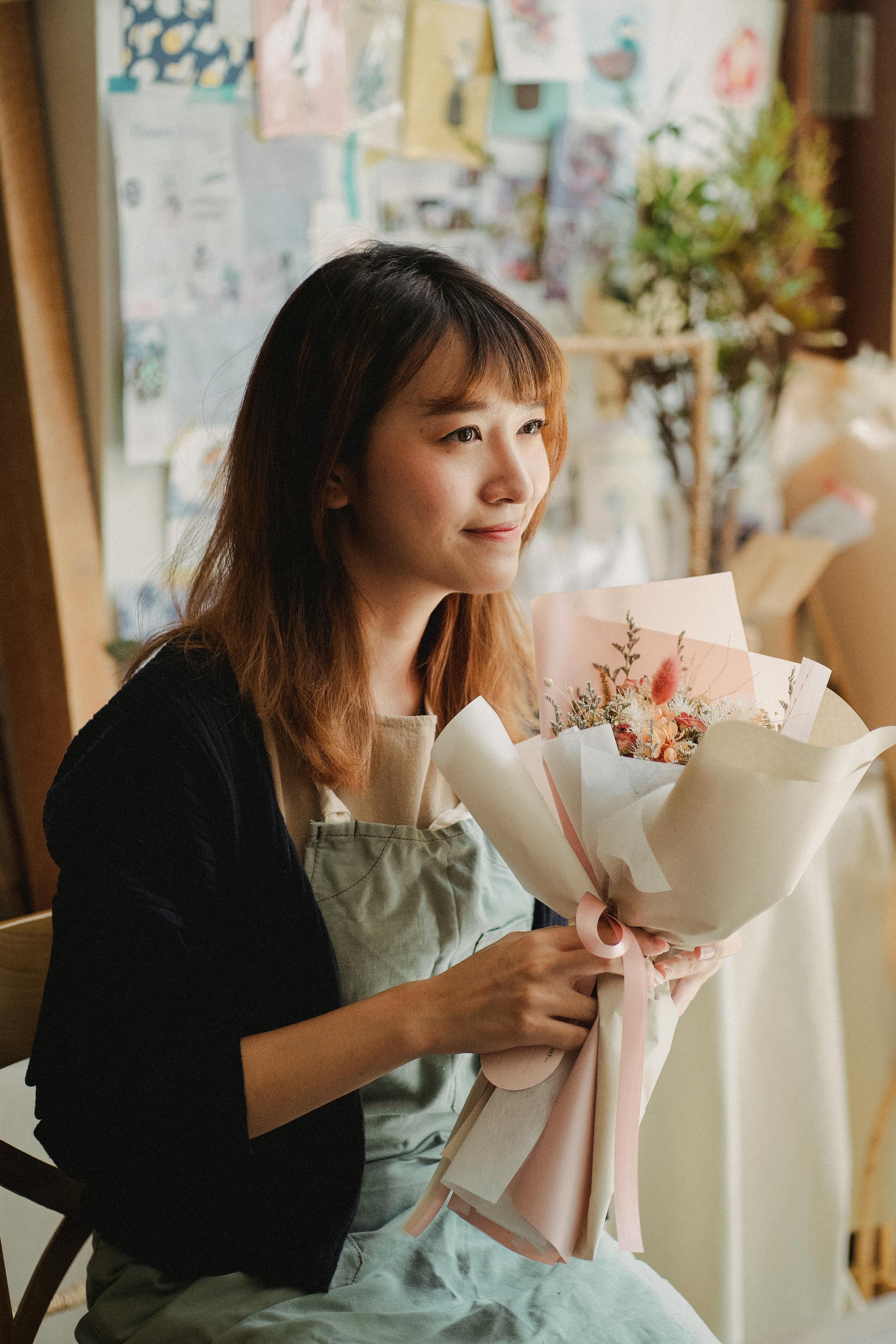 Friendly Asian female in employee apron looking away sitting on chair and holding pink bouquet of flowers