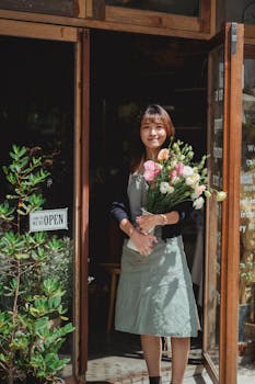 Young Asian female in apron looking at camera while holding bouquet of flowers standing at shop entrance