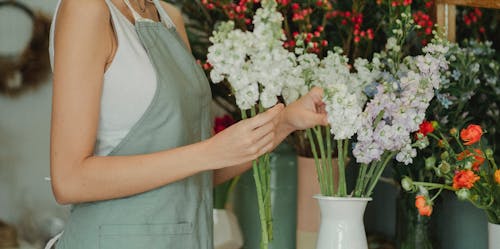 Crop anonymous florist in apron standing near table with flowers in vases and choosing flowers for composition
