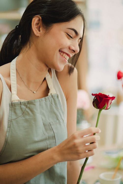Side view of happy ethnic female in casual clothes and apron holding fresh red rose and smiling with closed eyes in light room