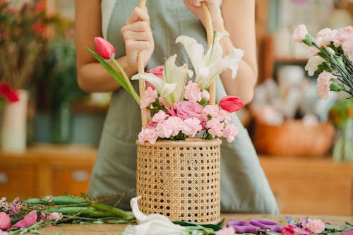 Crop anonymous female florist in light green apron standing with aromatic lily and tender pink tulips bouquet arranged in wicker basket in sunny floral shop