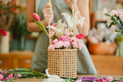 Crop anonymous female florist in light green apron standing with aromatic lily and tender pink tulips bouquet arranged in wicker basket in sunny floral shop