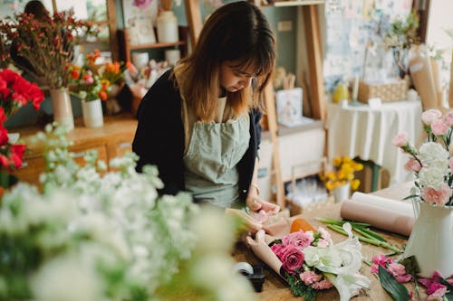 Concentrated young female florist in uniform standing near counter and arranging bouquet while working in light floral shop