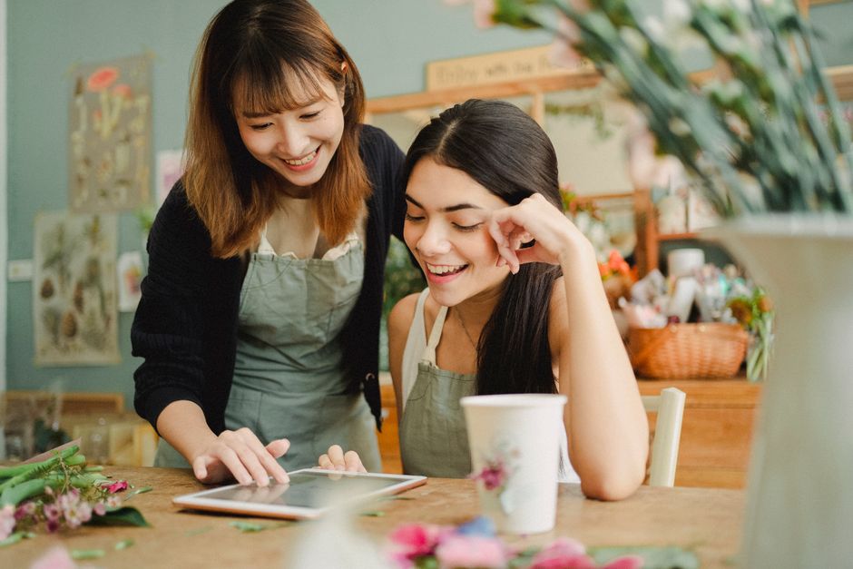 Smiling young diverse female floral shop coworkers wearing aprons surfing internet on modern tablet while working together