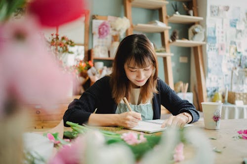 Glad Asian florist writing in notebook in flower shop