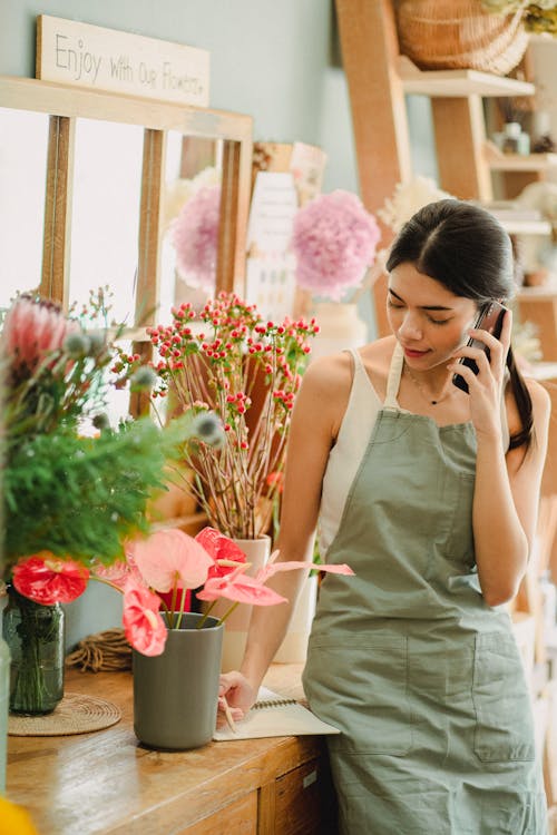 Free Focused young female florist in apron having conversation on mobile phone and taking notes in notebook while working in modern floristry studio Stock Photo