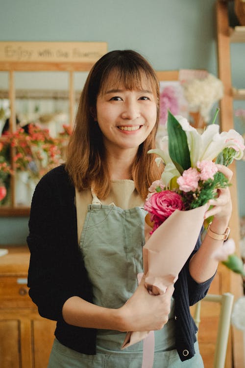 Attractive positive Asian female wearing green apron standing with wonderful bouquet in hands and looking at camera while working in light florists shop