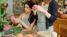 Positive young female florists wearing aprons removing leaves from flower stems before arranging bouquet in modern floristry store