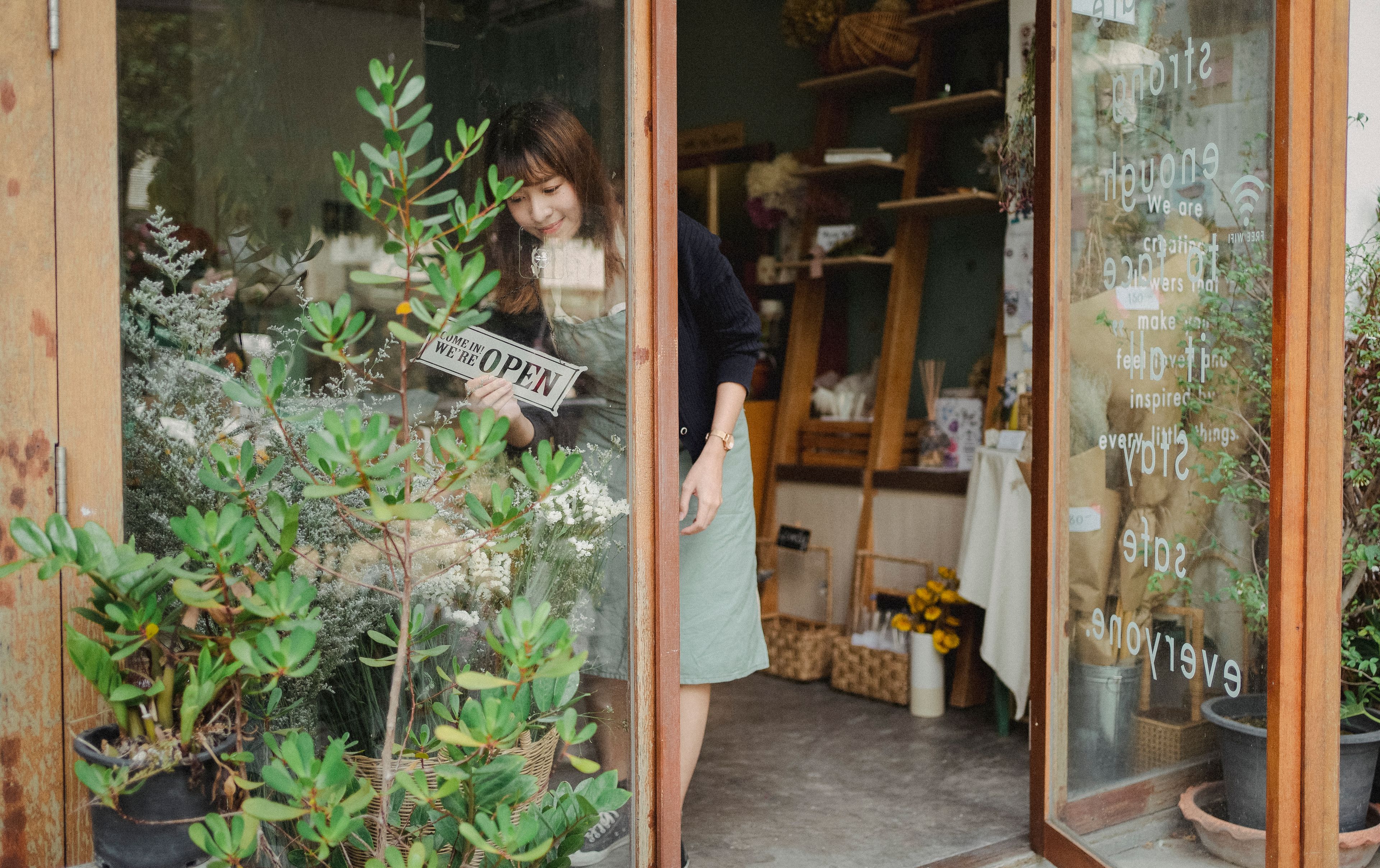 A shopkeeper turning the open sign in her plant-filled shop window