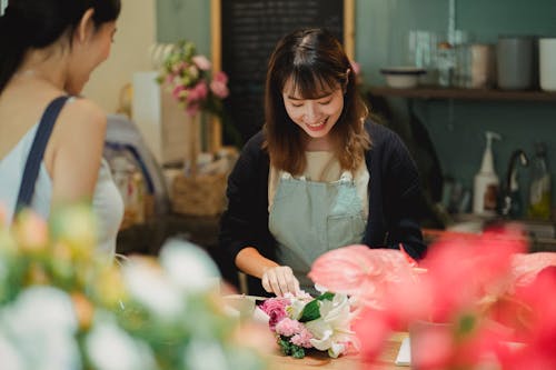 Content Asian female florist wearing light green apron composing tender bouquet for positive female customer while working at counter in floristry shop