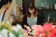 一位在花店柜台工作的亚洲女店员，身着深绿色围裙，为一位积极的女性顾客精心搭配了一束温柔的花束