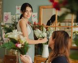 Side view glad young female florists wearing aprons arranging delicate flowers and looking at each other contentedly while working together in light floral store