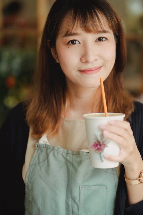 Crop glad young Asian female wearing light green apron enjoying refreshing drink from paper cup and looking at camera