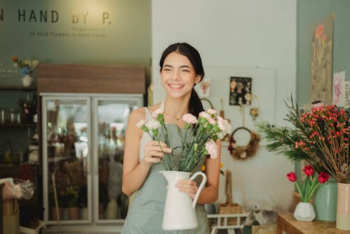 Happy young female florist wearing apron standing with tender pastel carnation flowers in white vase while working in floristry store and looking away with charming smile