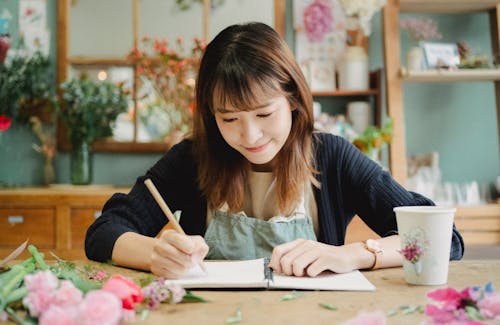 Glad young Asian female in uniform taking notes in notebook while working in light floral shop and sitting at table covered with many flowers petals