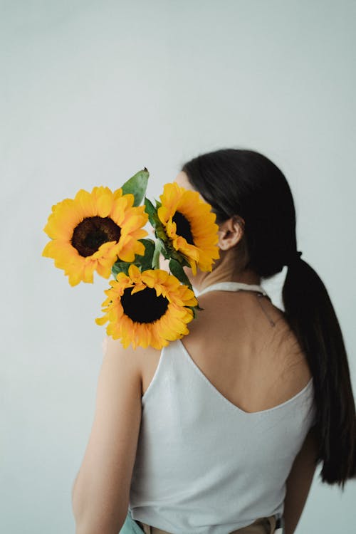 Back view of unrecognizable female with long dark hair in casual clothes standing with bunch of fresh sunflowers on shoulder against light background