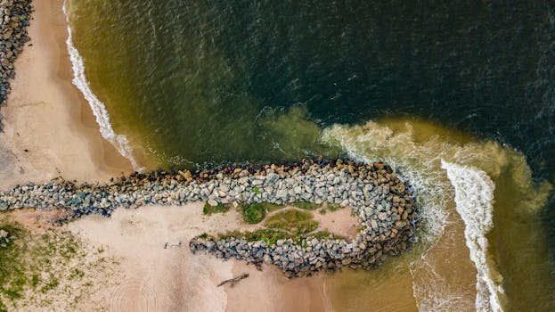 Drone shot capturing a rocky pier on a sandy beach with waves crashing against the shore.