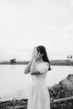 一张黑白照片中，一位身着时尚服装的女士在河岸上，以手遮面，天空多云