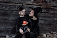 一对穿着女巫服装的母女拿着南瓜，靠在户外的木墙边