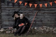 一位打扮成女巫的妇女和孩子面带微笑，坐在户外，手里拿着南瓜和万圣节装饰品