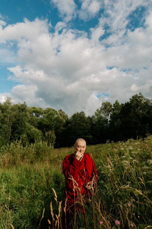 Free Monk in traditional robe standing in a tranquil meadow with lush greenery and cloudy sky. Stock Photo