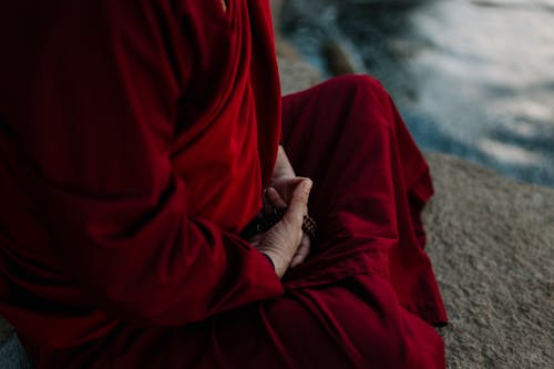 A Person Wearing a Red Robe Sitting on Rock with Prayer Beads