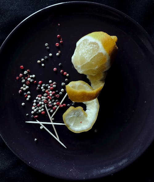 Free Top view composition of half peeled sour lemon with twisted zest placed on black plate with colorful peppercorns and toothpicks Stock Photo