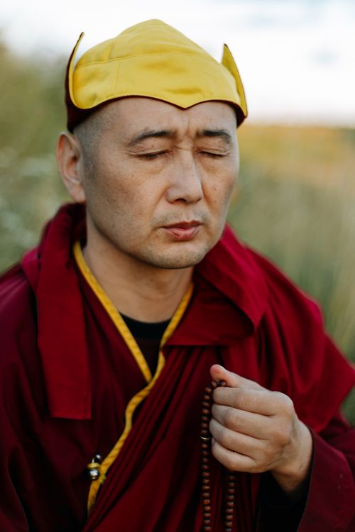 Free Tibetan monk in a red robe meditating outdoors with prayer beads, eyes closed for concentration. Stock Photo