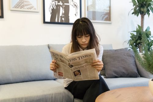 Free A young woman attentively reads a newspaper while sitting on a modern sofa indoors. Stock Photo