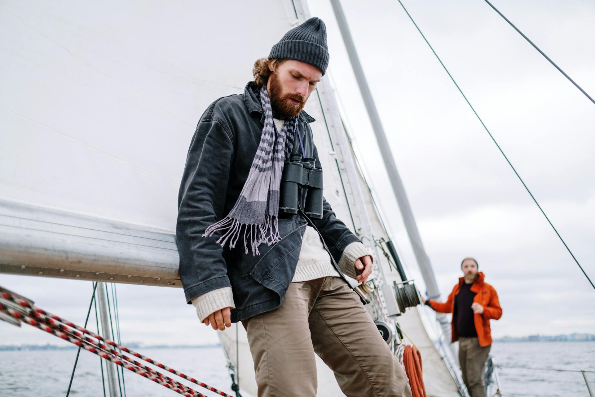 Two men sailing on a boat on a calm sea, dressed warmly, under cloudy skies.