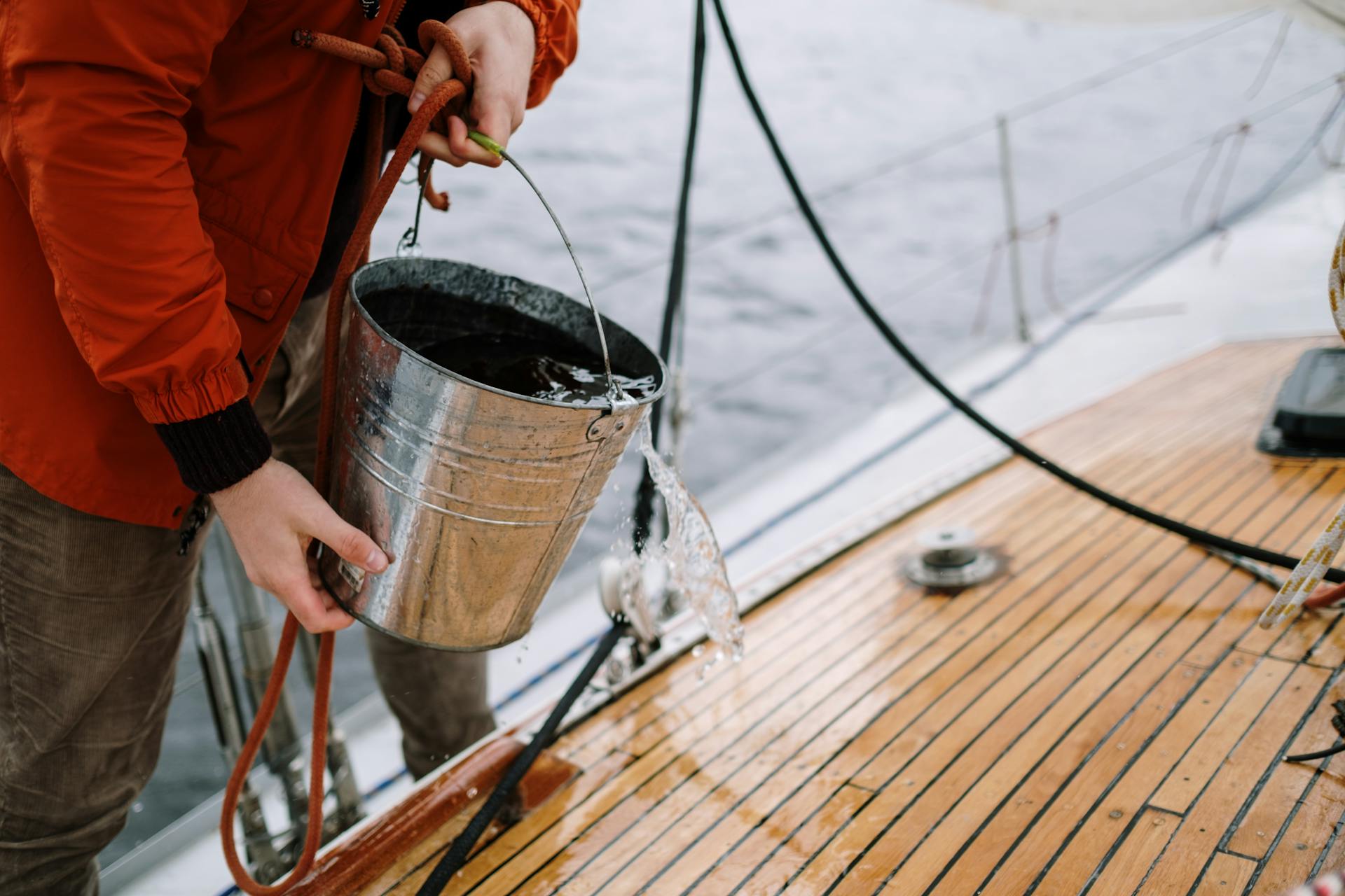 Close-up of a man washing a sailboat deck with water from a metal bucket outdoors.