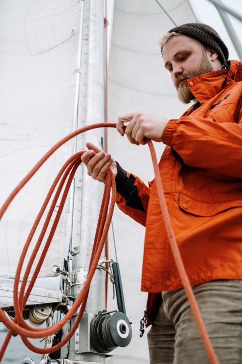 Free Adult male in orange jacket handling a sailing rope on a sailboat, depicting a nautical theme. Stock Photo