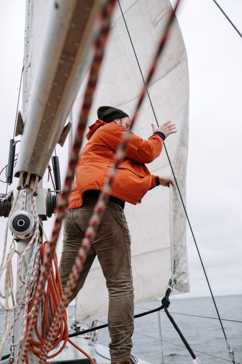 Free A man in an orange jacket adjusting sails on a sailboat during an ocean adventure. Stock Photo