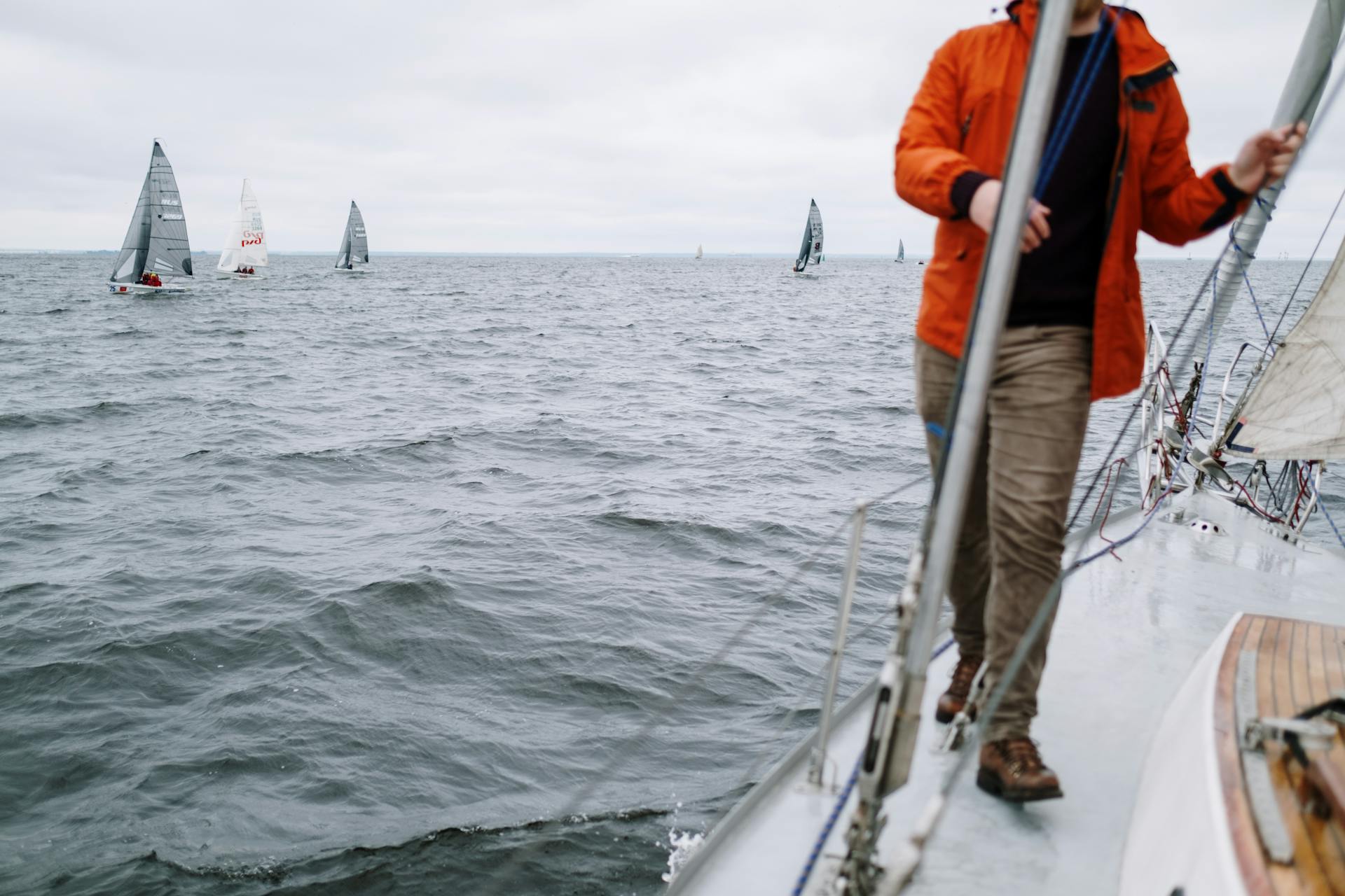 A man in an orange jacket sailing on a yacht observing a race of sailboats on the open sea.