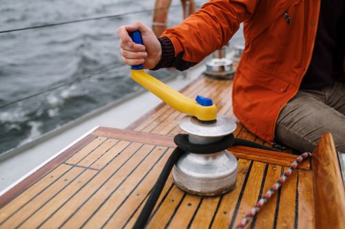Free Man in orange jacket operating winch on sailboat deck during daytime sailing adventure. Stock Photo