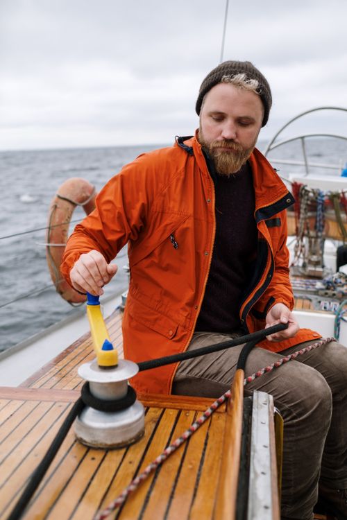 Free Adult man in orange jacket handling rope on a sailboat deck in overcast weather at sea. Stock Photo
