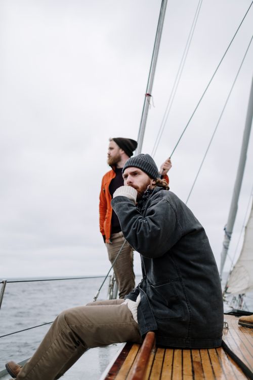 Free Two men sailing on a boat in the ocean under a gray overcast sky, wearing jackets and beanies. Stock Photo
