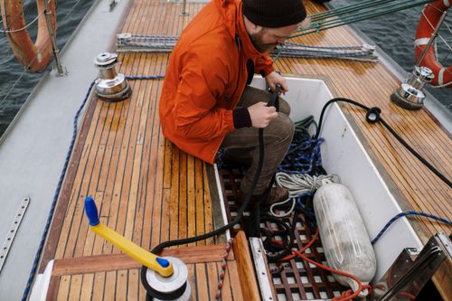 Free A man in an orange jacket tends to ropes and equipment on a wooden sailboat deck. Stock Photo