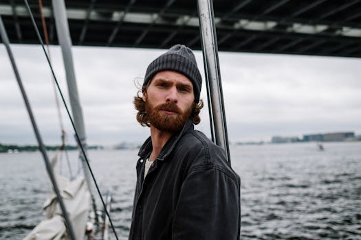 Bearded man with hat stands on a sailboat beneath a bridge, capturing a moody nautical vibe.