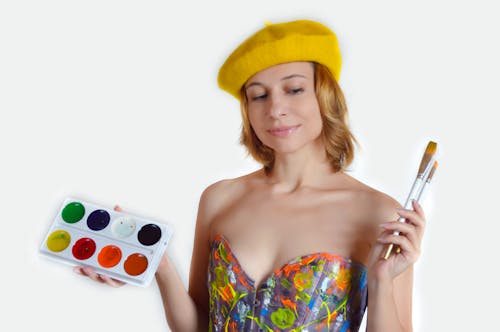 Thoughtful young female in colorful outfit and yellow beret with palette of watercolor and brushes standing against white background and looking down