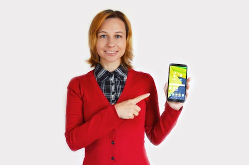 Cheerful young female in red cardigan and checkered shirt pointing at smartphone while standing against white background and looking at camera