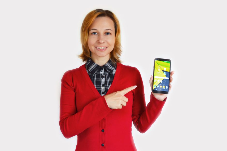 Cheerful young female in red cardigan and checkered shirt pointing at smartphone while standing against white background