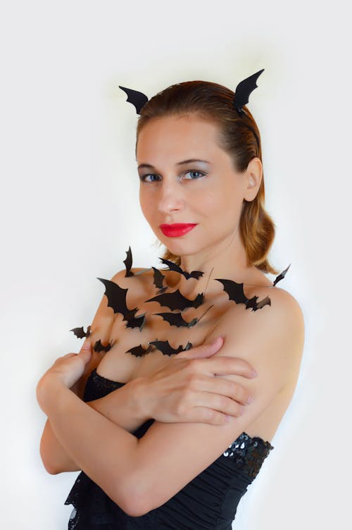 Free Young female in black dress decorated with small cutout bats for Halloween standing against white background and looking at camera Stock Photo