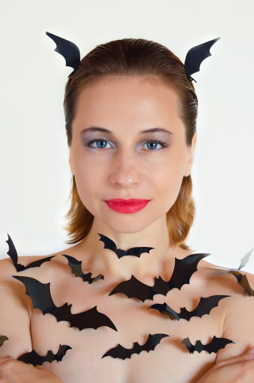 Free Naked young female wearing makeup and decorated with small black cutout bats for Halloween standing against white background and looking at camera Stock Photo