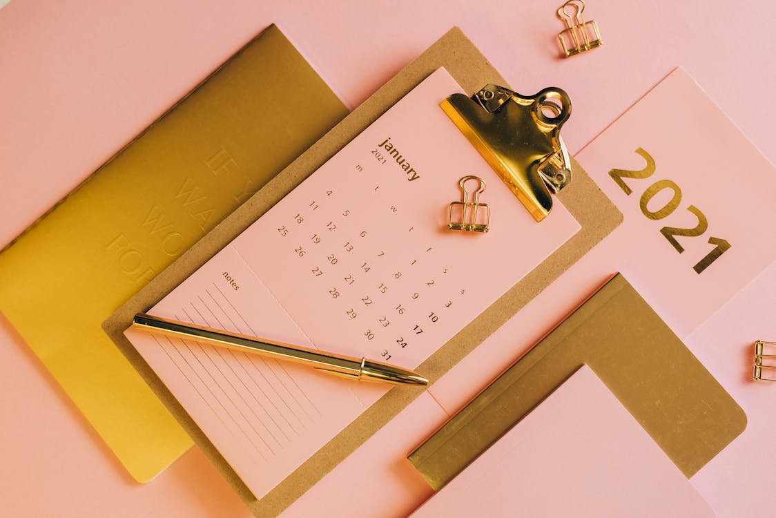 From above stylish workplace consisting of clipboard with calendar and golden notebook on pink background