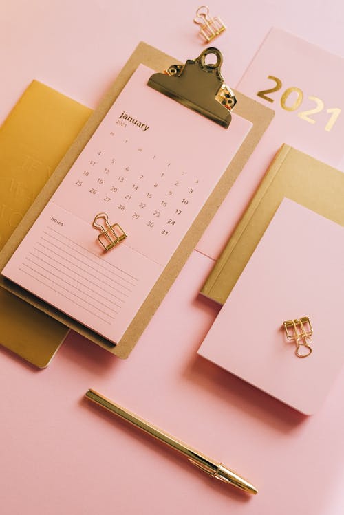 From above minimalistic composition of clipboard with calendar paper surrounded notebooks and clips on pink background